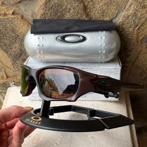 Może przedstawiać: Brązowe okulary przeciwsłoneczne Oakley z odblaskowymi soczewkami, prezentowane na czarnym stojaku z logo Oakley. Srebrne etui i czarna ściereczka znajdują się za okularami. Tło to kamienna ściana.