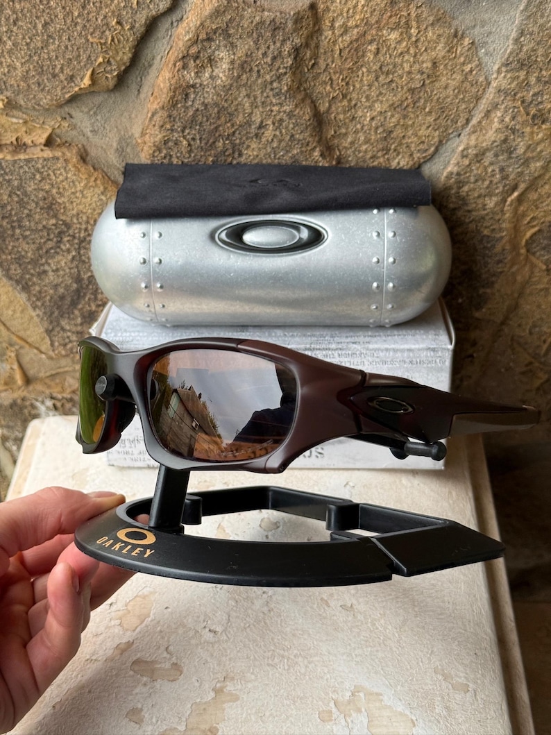 Może przedstawiać: Brązowe okulary przeciwsłoneczne Oakley z odblaskowymi soczewkami, prezentowane na czarnym stojaku z logo Oakley. Srebrne etui i czarna ściereczka znajdują się za okularami. Tło to kamienna ściana.