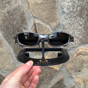以下が含まれることがあります： 黒いレンズのシルバーフレームのOakleyサングラスが黒いスタンドに展示されています。スタンドには金色の文字でOakleyのロゴが入っています。サングラスは、石壁を背景に手で持たれています。