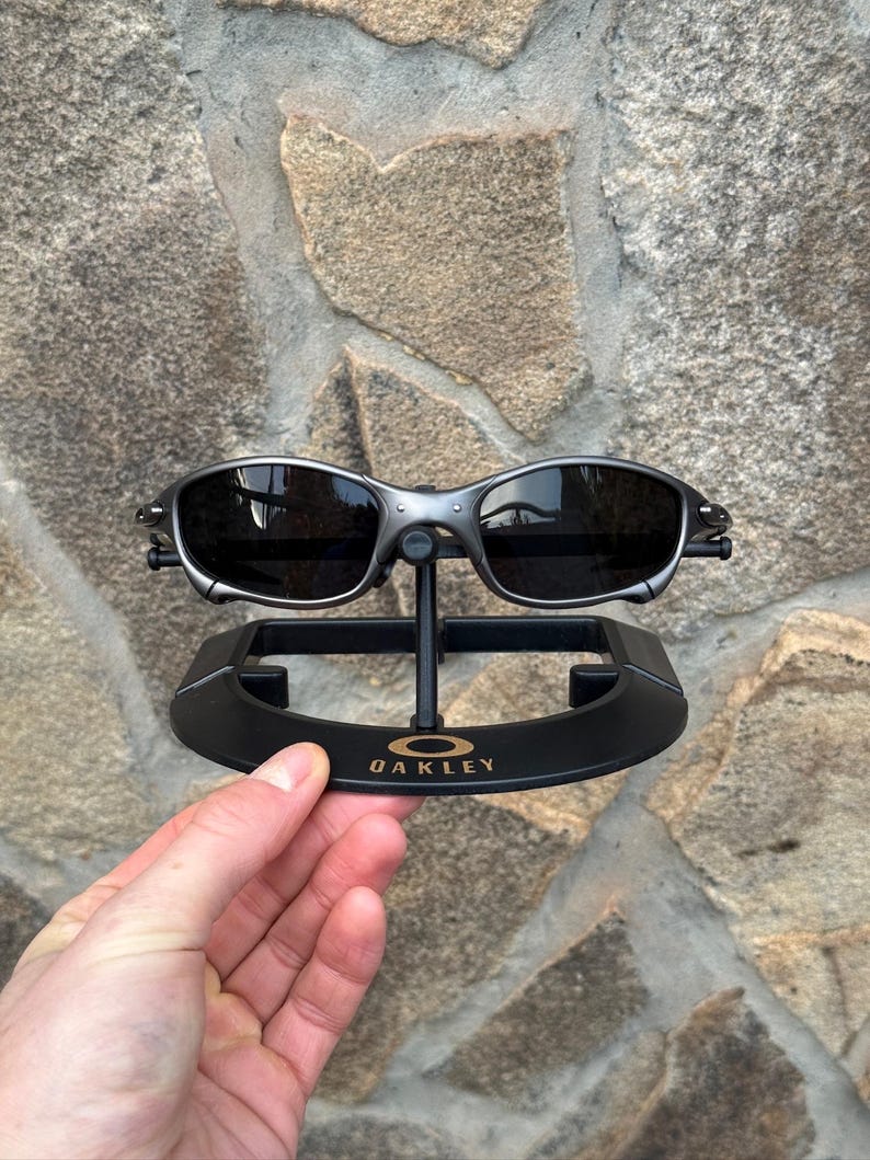 以下が含まれることがあります： 黒いレンズのシルバーフレームのOakleyサングラスが黒いスタンドに展示されています。スタンドには金色の文字でOakleyのロゴが入っています。サングラスは、石壁を背景に手で持たれています。