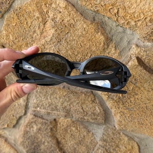 K&ouml;nnte beinhalten: Schwarze Sonnenbrille mit dunklen Gl&auml;sern. Die Sonnenbrille hat ein schlankes, modernes Design mit matter Oberfl&auml;che. Die B&uuml;gel der Sonnenbrille sind d&uuml;nn und leicht gebogen. Das Oakley-Logo ist auf dem Rahmen sichtbar.