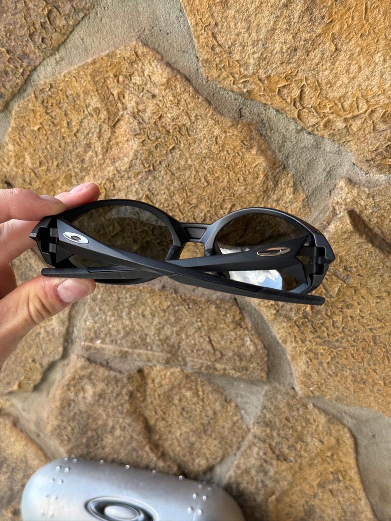 K&ouml;nnte beinhalten: Schwarze Sonnenbrille mit dunklen Gl&auml;sern. Die Sonnenbrille hat ein schlankes, modernes Design mit matter Oberfl&auml;che. Die B&uuml;gel der Sonnenbrille sind d&uuml;nn und leicht gebogen. Das Oakley-Logo ist auf dem Rahmen sichtbar.