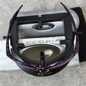 Puede incluir: Gafas de sol Oakley negras y moradas con un patr&oacute;n moteado en las patillas, apoyadas en un expositor negro. Las lentes son de color morado claro. El expositor tiene el logotipo de Oakley y el texto "SCIENCE AND ART".