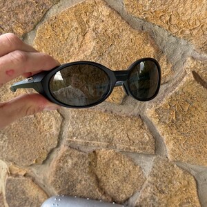 K&ouml;nnte beinhalten: Schwarze Sonnenbrille mit ovalen Gl&auml;sern und einem passenden schwarzen Rahmen. Die Sonnenbrille wird vor einer Steinmauer gehalten. Ein silbernes Etui mit dem Oakley-Logo ist darunter zu sehen.