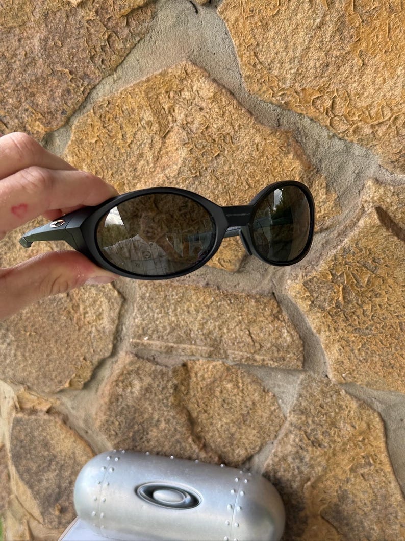 K&ouml;nnte beinhalten: Schwarze Sonnenbrille mit ovalen Gl&auml;sern und einem passenden schwarzen Rahmen. Die Sonnenbrille wird vor einer Steinmauer gehalten. Ein silbernes Etui mit dem Oakley-Logo ist darunter zu sehen.