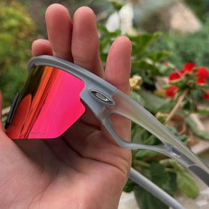 May include: Gray sunglasses with red-tinted lenses. The sunglasses have a modern design with a gray frame and a silver logo on the side. The lenses are a vibrant red color, providing a bold contrast to the gray frame.