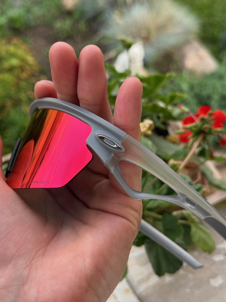 May include: Gray sunglasses with red-tinted lenses. The sunglasses have a modern design with a gray frame and a silver logo on the side. The lenses are a vibrant red color, providing a bold contrast to the gray frame.