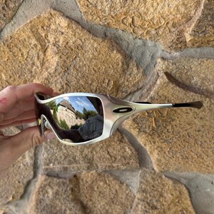 May include: Silver-framed sunglasses with mirrored lenses. The sunglasses have a sleek, modern design with a curved frame and dark temples. The lenses reflect the surrounding environment. The sunglasses are held against a textured stone background.