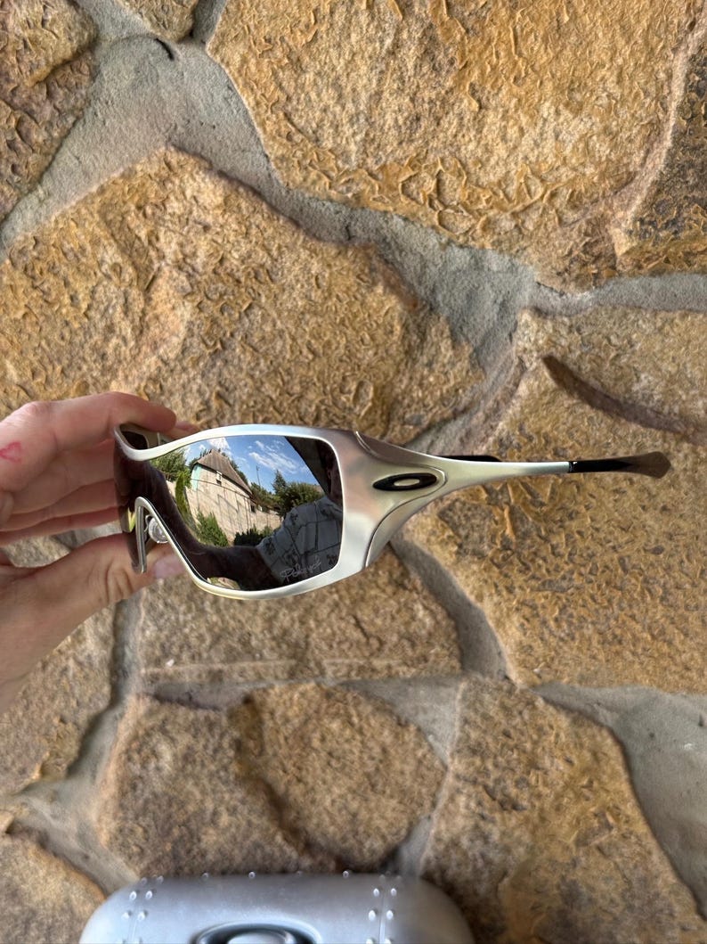 May include: Silver-framed sunglasses with mirrored lenses. The sunglasses have a sleek, modern design with a curved frame and dark temples. The lenses reflect the surrounding environment. The sunglasses are held against a textured stone background.