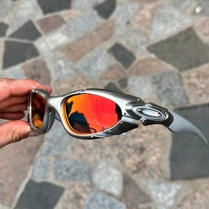 以下が含まれることがあります： 鮮やかな赤とオレンジ色のレンズが付いたシルバーフレームのサングラス。 サングラスは、湾曲したテンプルアームを備えた洗練されたモダンなデザインです。 レンズは周囲の環境を反射し、ダイナミックな視覚効果を生み出します。 フレームはメタリックシルバーです。