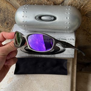 May include: A pair of Oakley sunglasses with a metallic gray frame and purple lenses. The sunglasses are displayed with a matching silver case and a black microfiber pouch. The case has a silver Oakley logo.