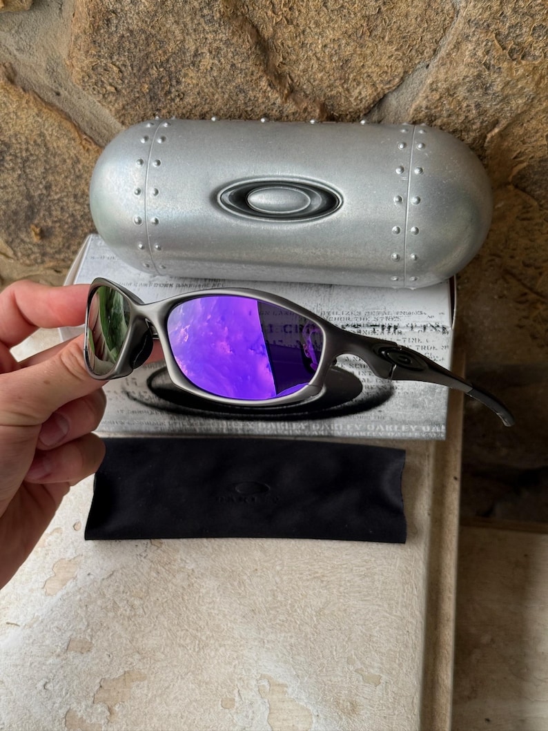 May include: A pair of Oakley sunglasses with a metallic gray frame and purple lenses. The sunglasses are displayed with a matching silver case and a black microfiber pouch. The case has a silver Oakley logo.