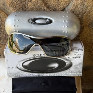 May include: Silver Oakley sunglasses with a black lens, resting on a white box with the word "SCIENCE" printed on it. A silver case and a black microfiber cloth are also present. The sunglasses have a metallic frame.