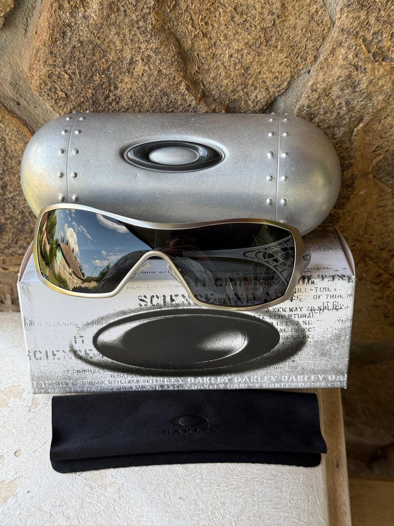 May include: Silver Oakley sunglasses with a black lens, resting on a white box with the word "SCIENCE" printed on it. A silver case and a black microfiber cloth are also present. The sunglasses have a metallic frame.