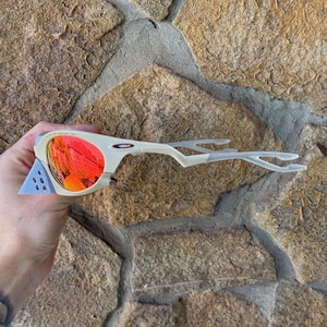 May include: Off-white and light blue sunglasses with red-orange lenses. The sunglasses have a unique, angular design with a distinctive frame and temples. The lenses reflect a vibrant, fiery color. The sunglasses are a sporty accessory.