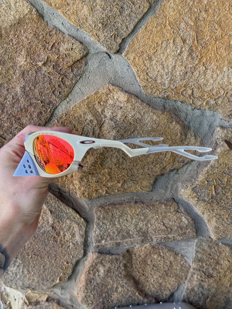 May include: Off-white and light blue sunglasses with red-orange lenses. The sunglasses have a unique, angular design with a distinctive frame and temples. The lenses reflect a vibrant, fiery color. The sunglasses are a sporty accessory.
