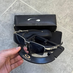 May include: Oakley sunglasses with a silver frame and dark lenses. The sunglasses are displayed with a black case and a black display stand with the Oakley logo in gold. The sunglasses are rectangular.
