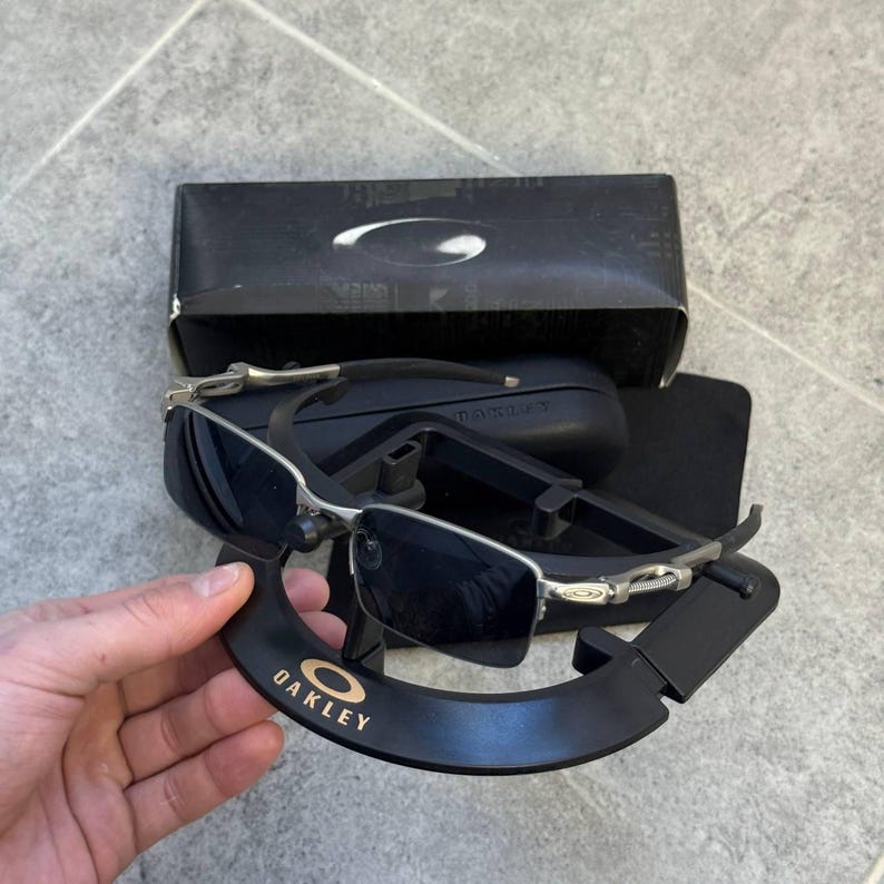 May include: Oakley sunglasses with a silver frame and dark lenses. The sunglasses are displayed with a black case and a black display stand with the Oakley logo in gold. The sunglasses are rectangular.