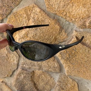 K&ouml;nnte beinhalten: Schwarze Sonnenbrille mit geschwungenem Rahmen und dunklen Gl&auml;sern. Die Sonnenbrille hat ein kleines silbernes Logo an der Seite und eine rot-wei&szlig;e USA-Markierung am B&uuml;gel. Die Gl&auml;ser spiegeln die Umgebung wider.