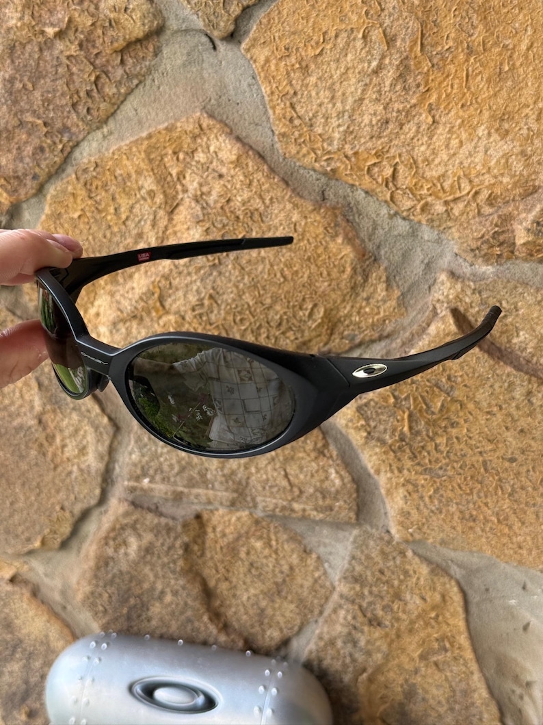 K&ouml;nnte beinhalten: Schwarze Sonnenbrille mit geschwungenem Rahmen und dunklen Gl&auml;sern. Die Sonnenbrille hat ein kleines silbernes Logo an der Seite und eine rot-wei&szlig;e USA-Markierung am B&uuml;gel. Die Gl&auml;ser spiegeln die Umgebung wider.