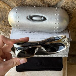 May include: Silver-framed sunglasses with dark lenses, a black case, and a silver case with the Oakley logo. The sunglasses have a unique design with intricate detailing on the temples. The case is oval-shaped with rivets.