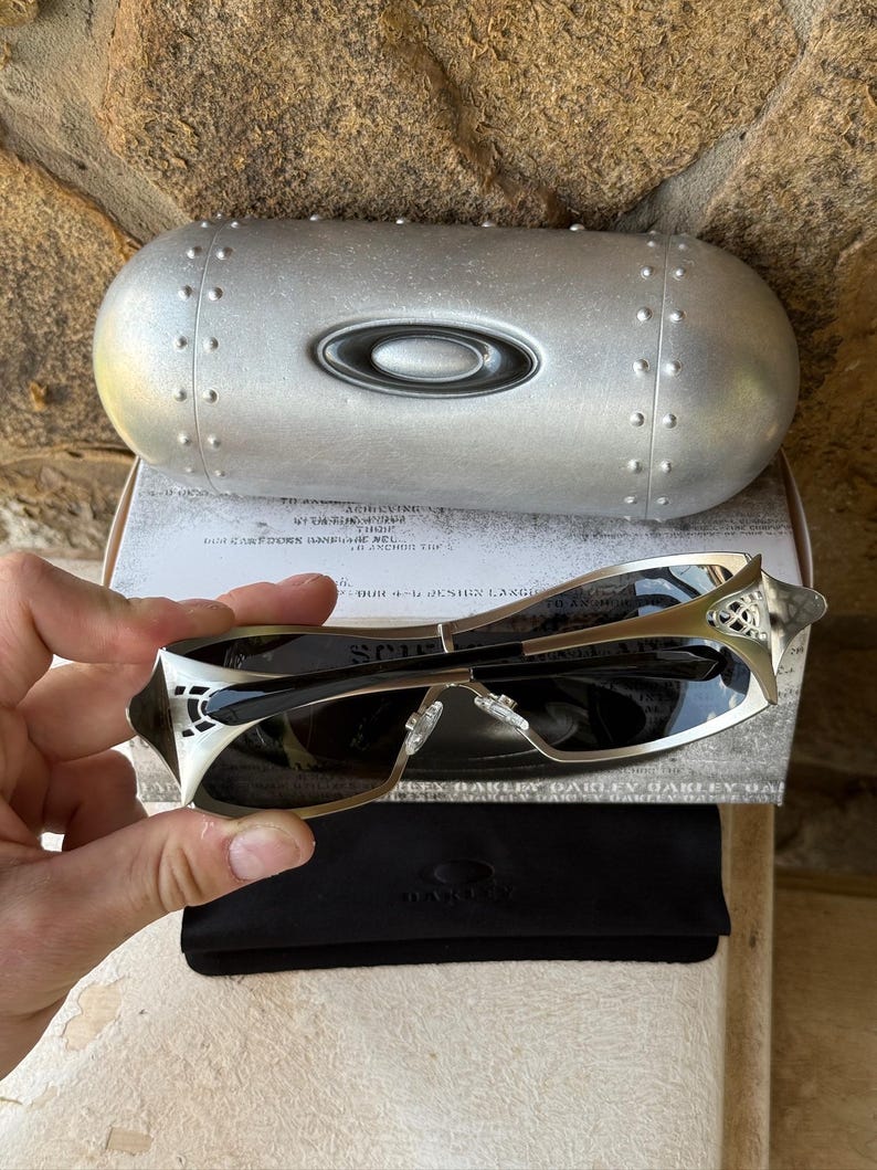 May include: Silver-framed sunglasses with dark lenses, a black case, and a silver case with the Oakley logo. The sunglasses have a unique design with intricate detailing on the temples. The case is oval-shaped with rivets.