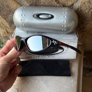 May include: Black Oakley sunglasses with mirrored lenses, a white star design, and a silver case. The case has a riveted design and the Oakley logo. A black microfiber bag and a box with text are also present.