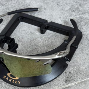 May include: Silver-framed sunglasses with green-tinted lenses, resting on a black display stand. The Oakley logo is visible on the frame. The sunglasses have a sleek, modern design.