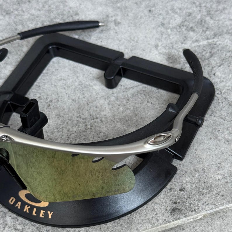 May include: Silver-framed sunglasses with green-tinted lenses, resting on a black display stand. The Oakley logo is visible on the frame. The sunglasses have a sleek, modern design.