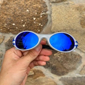 May include: Silver-framed sunglasses with vibrant blue lenses. The sunglasses have a sporty design with white and blue accents on the temples. The lenses reflect the surrounding environment.