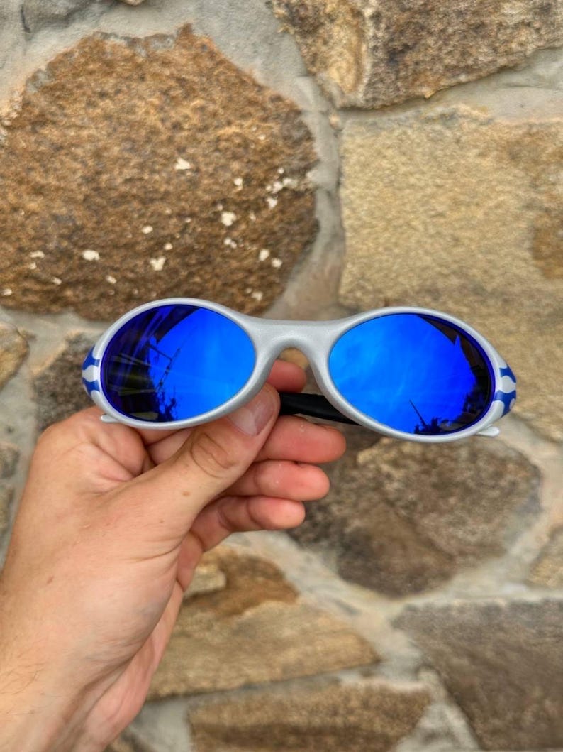 May include: Silver-framed sunglasses with vibrant blue lenses. The sunglasses have a sporty design with white and blue accents on the temples. The lenses reflect the surrounding environment.