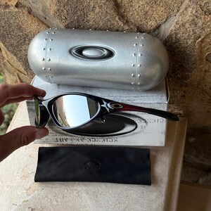 May include: Black and silver sunglasses with a reflective lens, resting on a white box with the text "SCIENCE AND ART". A silver case and a black microfiber cloth are also present. The sunglasses have a sleek, modern design.