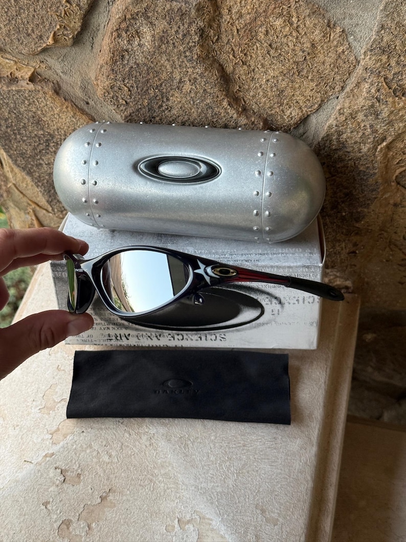 May include: Black and silver sunglasses with a reflective lens, resting on a white box with the text "SCIENCE AND ART". A silver case and a black microfiber cloth are also present. The sunglasses have a sleek, modern design.
