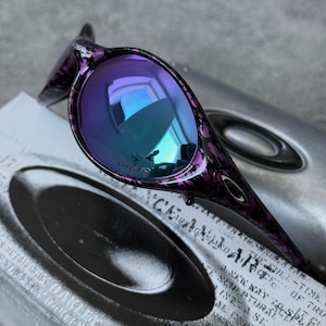 Puede incluir: Gafas de sol Oakley moradas y negras con lentes iridiscentes azules y moradas. La montura tiene un dise&ntilde;o estampado. Las gafas de sol est&aacute;n sobre un estuche plateado con el logotipo y texto de Oakley.