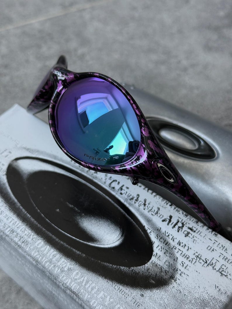 Puede incluir: Gafas de sol Oakley moradas y negras con lentes iridiscentes azules y moradas. La montura tiene un dise&ntilde;o estampado. Las gafas de sol est&aacute;n sobre un estuche plateado con el logotipo y texto de Oakley.