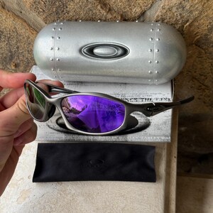 May include: Silver-framed sunglasses with purple mirrored lenses, a black microfiber pouch, and a silver case. The case has a riveted design and the Oakley logo. The sunglasses are held in a hand.