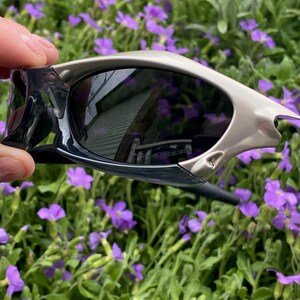 May include: Sport sunglasses with a silver frame and dark lenses. The sunglasses have a sleek, modern design with a curved shape. The lenses reflect the surrounding environment. The sunglasses are held against a backdrop of green foliage and purple flowers.