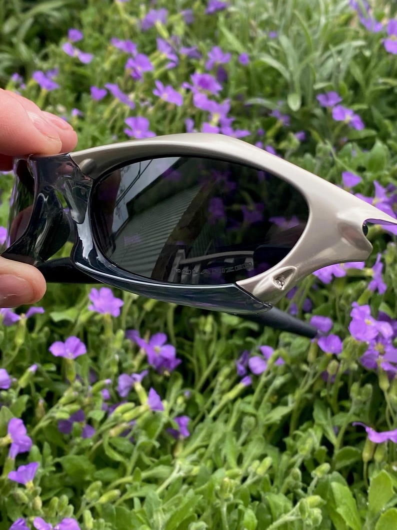 May include: Sport sunglasses with a silver frame and dark lenses. The sunglasses have a sleek, modern design with a curved shape. The lenses reflect the surrounding environment. The sunglasses are held against a backdrop of green foliage and purple flowers.