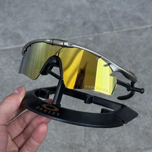 May include: Silver-framed sunglasses with yellow-tinted lenses, resting on a black display stand. The lenses reflect a golden hue, and the frame has a sleek, modern design. The text "MAGNESIUM" is visible on the lens.