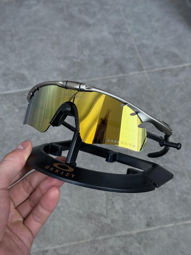 May include: Silver-framed sunglasses with yellow-tinted lenses, resting on a black display stand. The lenses reflect a golden hue, and the frame has a sleek, modern design. The text "MAGNESIUM" is visible on the lens.