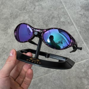 Puede incluir: Gafas de sol Oakley moradas y negras con lentes reflectantes. La montura tiene un dise&ntilde;o moteado y el texto "Brain Dead" es visible en la parte superior de la montura. Las gafas de sol se exhiben en un soporte negro con el logotipo de Oakley.