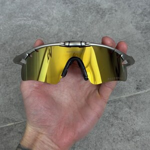 May include: Silver-framed sunglasses with mirrored gold lenses. The sunglasses have a black nose bridge and are held in a hand. The lenses reflect the surrounding environment. The brand name is visible on the lens.