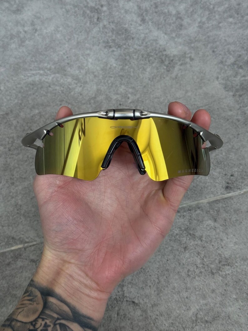 May include: Silver-framed sunglasses with mirrored gold lenses. The sunglasses have a black nose bridge and are held in a hand. The lenses reflect the surrounding environment. The brand name is visible on the lens.