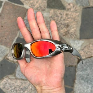 以下が含まれることがあります： 赤と黄色のレンズが付いたシルバーフレームのサングラス。 サングラスは、湾曲したテンプルアームを備えたユニークで角張ったデザインです。 レンズは鮮やかなオレンジ色を反射し、フレームはメタリック仕上げです。