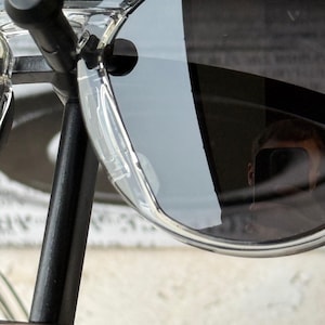 May include: Clear-framed sunglasses with dark lenses. The sunglasses are mounted on a black metal stand. The lenses reflect the surrounding environment. The frame is transparent and has a curved design.