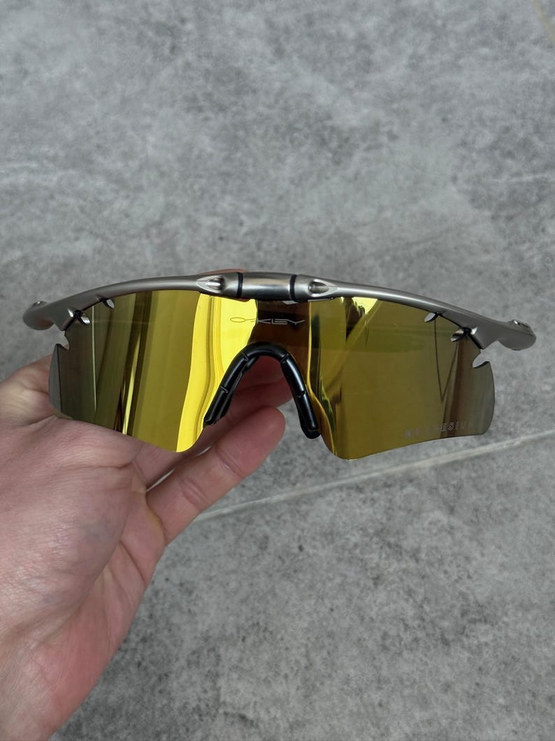 May include: Silver-framed sunglasses with gold-tinted lenses. The sunglasses have a black nose bridge and ear pieces. The lenses are a bright gold color, and the frame is a metallic silver.