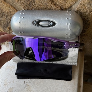 Könnte beinhalten: Violett-schwarze Oakley-Sonnenbrille mit verspiegelten Gläsern, einem silbernen Etui und einem schwarzen Mikrofasertuch. Die Sonnenbrille wird auf einer weißen Oberfläche mit Text präsentiert.