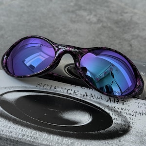 Puede incluir: Gafas de sol ovaladas con lentes moradas y montura moteada negra y morada. El texto "DRAW DEAD" es visible en la montura. Las lentes reflejan un tono azul y verde. Las gafas de sol est&aacute;n sobre una superficie impresa en blanco y negro.