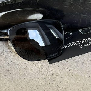 May include: Black rectangular sunglasses with dark lenses and a thin metal frame. The sunglasses are resting on a textured surface, with a black card visible behind them. The card has white text and a drawing of glasses.