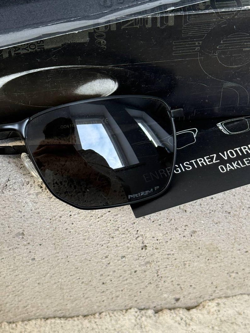 May include: Black rectangular sunglasses with dark lenses and a thin metal frame. The sunglasses are resting on a textured surface, with a black card visible behind them. The card has white text and a drawing of glasses.